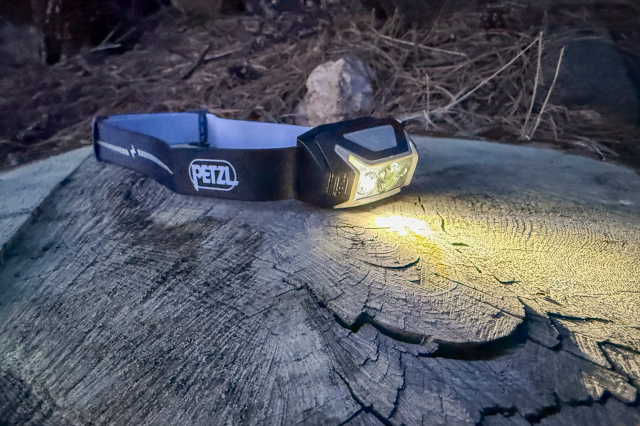 Petzl headlamp resting on a weathered log at dusk, shining a bright white beam across the wood grain.
