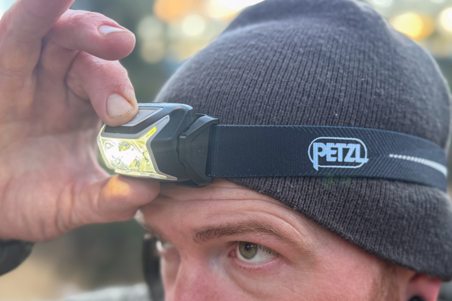 Side close-up of a person wearing a Petzl headlamp over a beanie while adjusting the light with one hand.