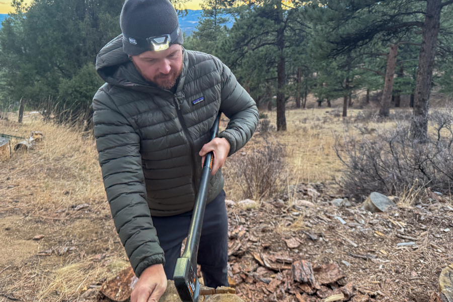 Person wearing a headlamp while handling firewood, outdoors on a dry hillside with trees in the background.