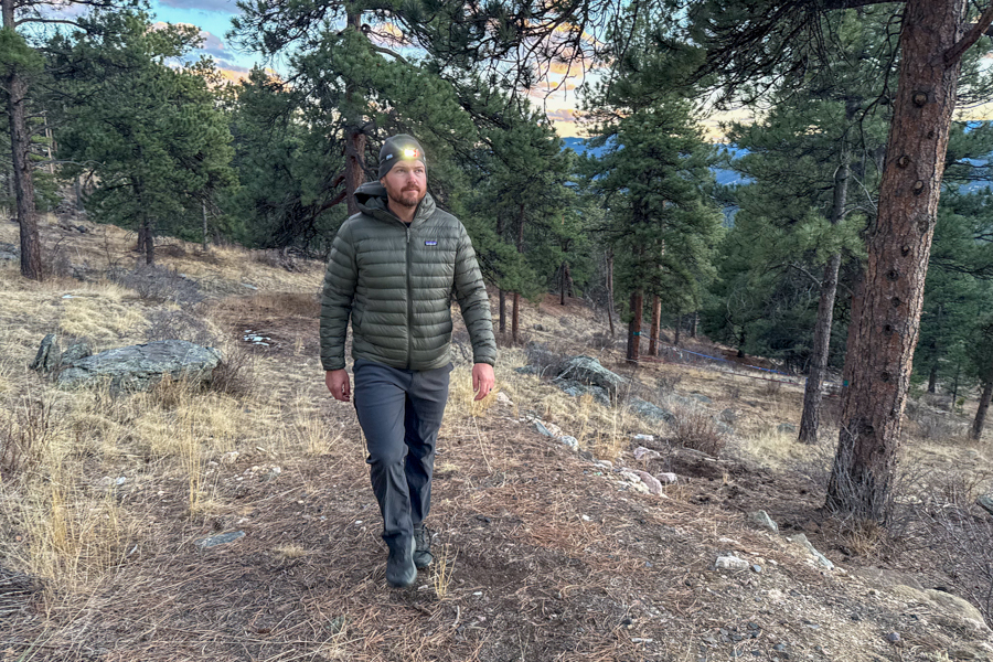 Person walking on a pine-covered trail at dusk with a headlamp shining on their forehead.