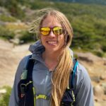 A woman with sunglasses stands in front of snow-covered mountains on a sunny day