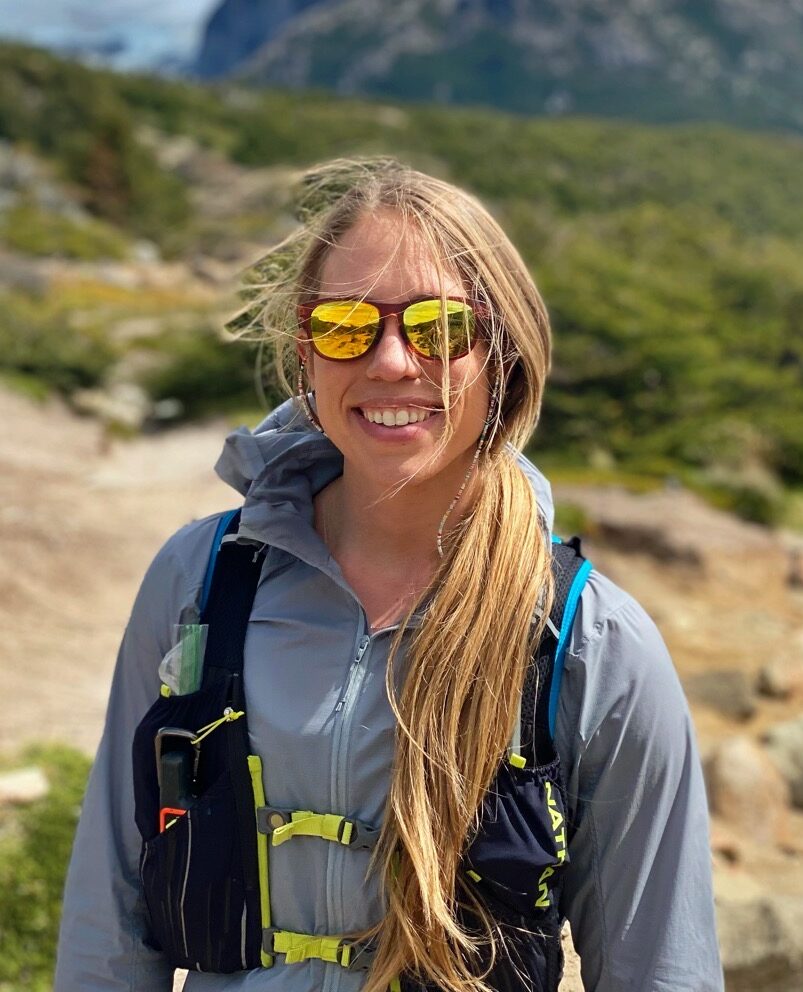 A woman with sunglasses stands in front of snow-covered mountains on a sunny day