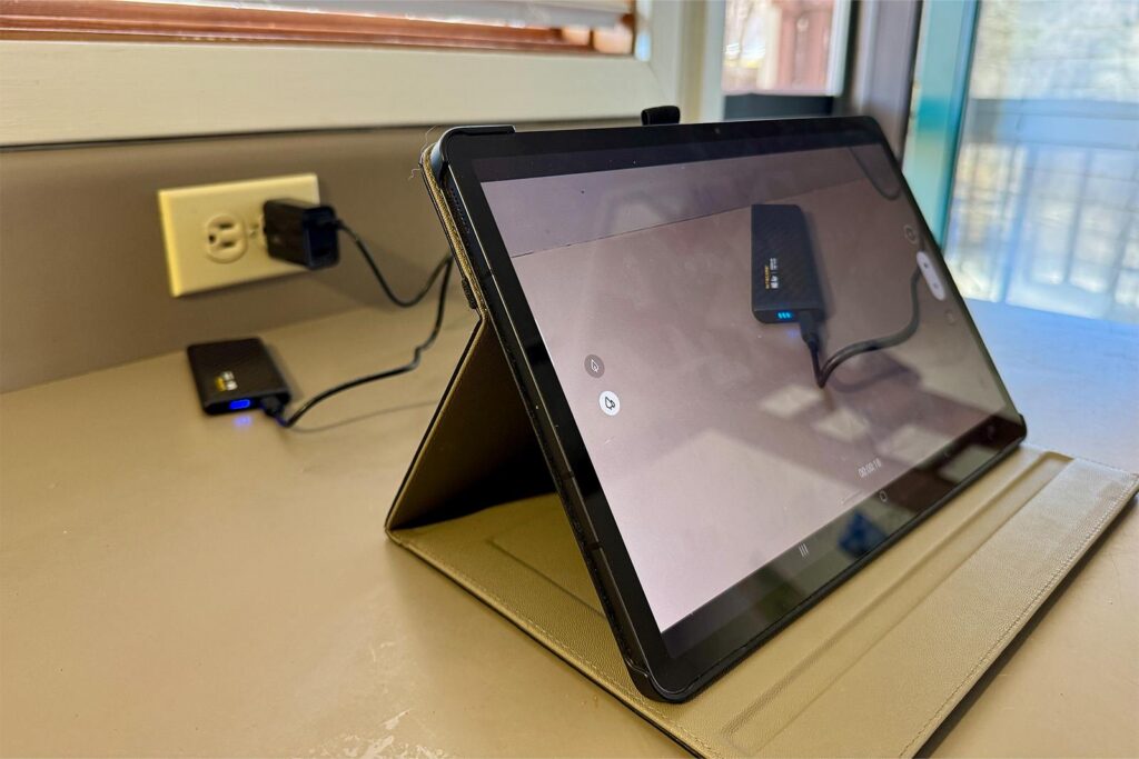 The Nitecore NB Air is plugged into a wall charger and leaning against a wall on a counter. In the foreground, a tablet is recording the NB Air.