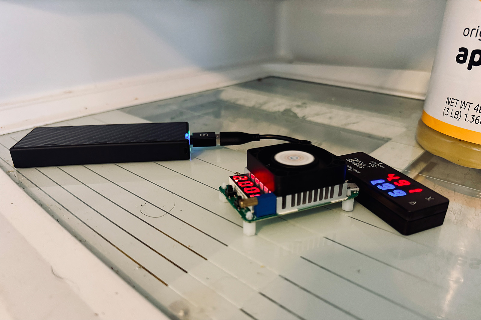 El banco de energía Nitecore NB10000 Gen 4 está conectado a un dispositivo multímetro digital. También hay una resistencia de carga ficticia conectada a él. Los tres están sentados en el estante inferior de una nevera con un poco de salsa de manzana.