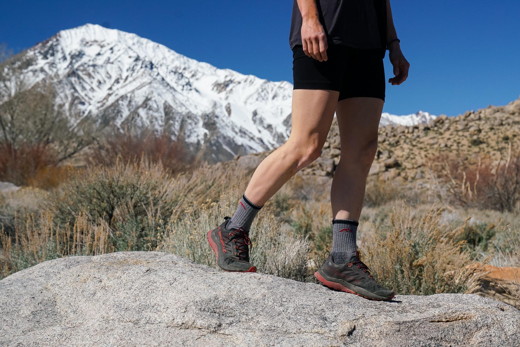 The bottom half of a person walks on top of a rock. They are wearing hiking socks and sneakers. There is a snow topped mountain in the background.