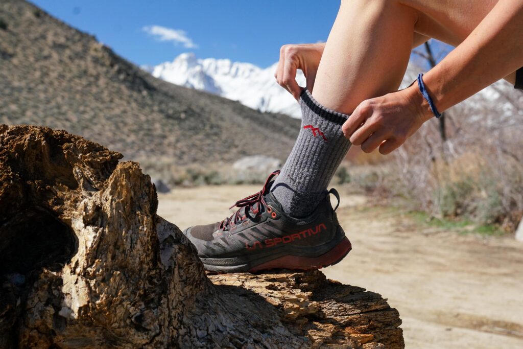 A close up image of a person pulling up a grey hiking sock around their leg. Their foot rests on a rock with mountains in the background.