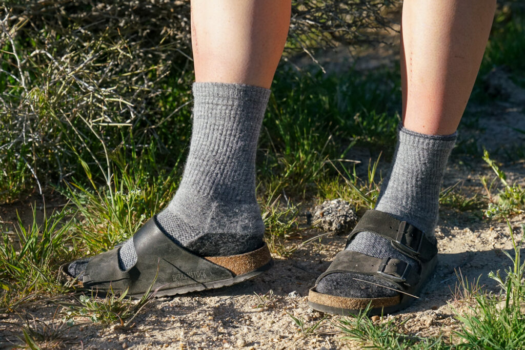 A close up image of the bottom half of a person's legs. They are wearing grey socks and sandals and stand in the grass.