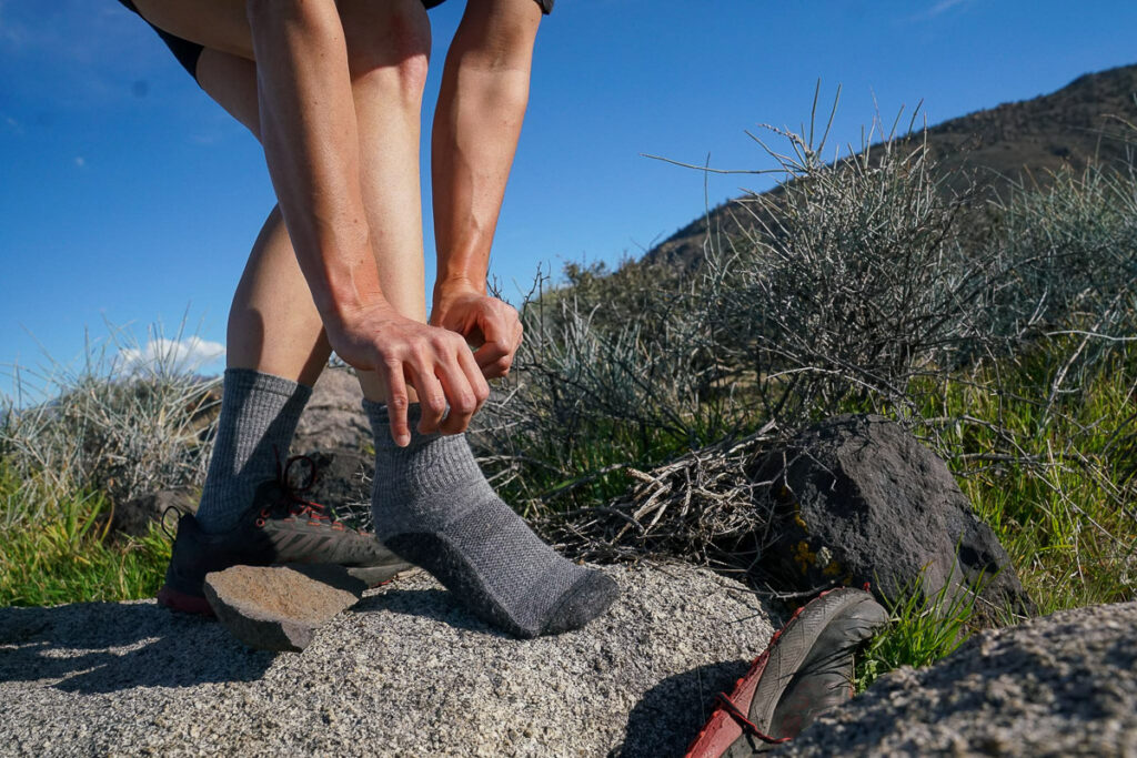 An image of the bottom half of a person adjusting a grey sock on their foot. They stand on a rock with bushes in the background.