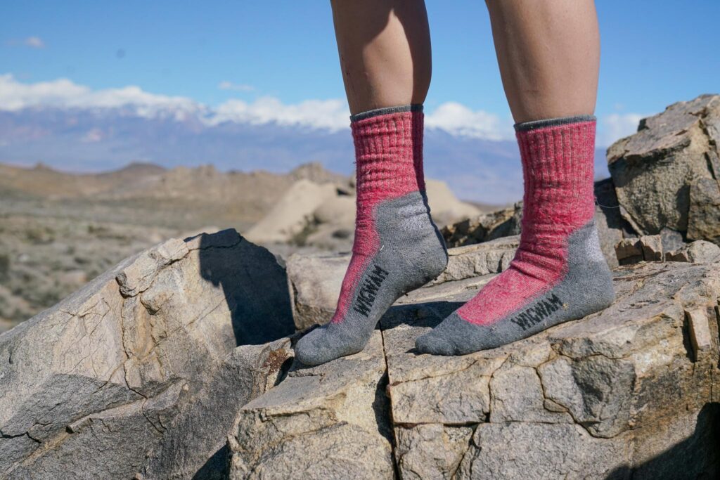 A close up image of a person's feet. They are wearing pink and grey hiking socks and stand on a rock with mountains in the background.