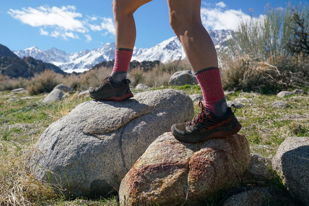 A close up image of a person's legs. They are standing on two rocks wearing sneakers and hiking socks. There are mountains in the background.
