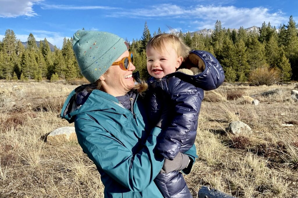 A smiling woman in a jacket holds a smiling baby in a jacket