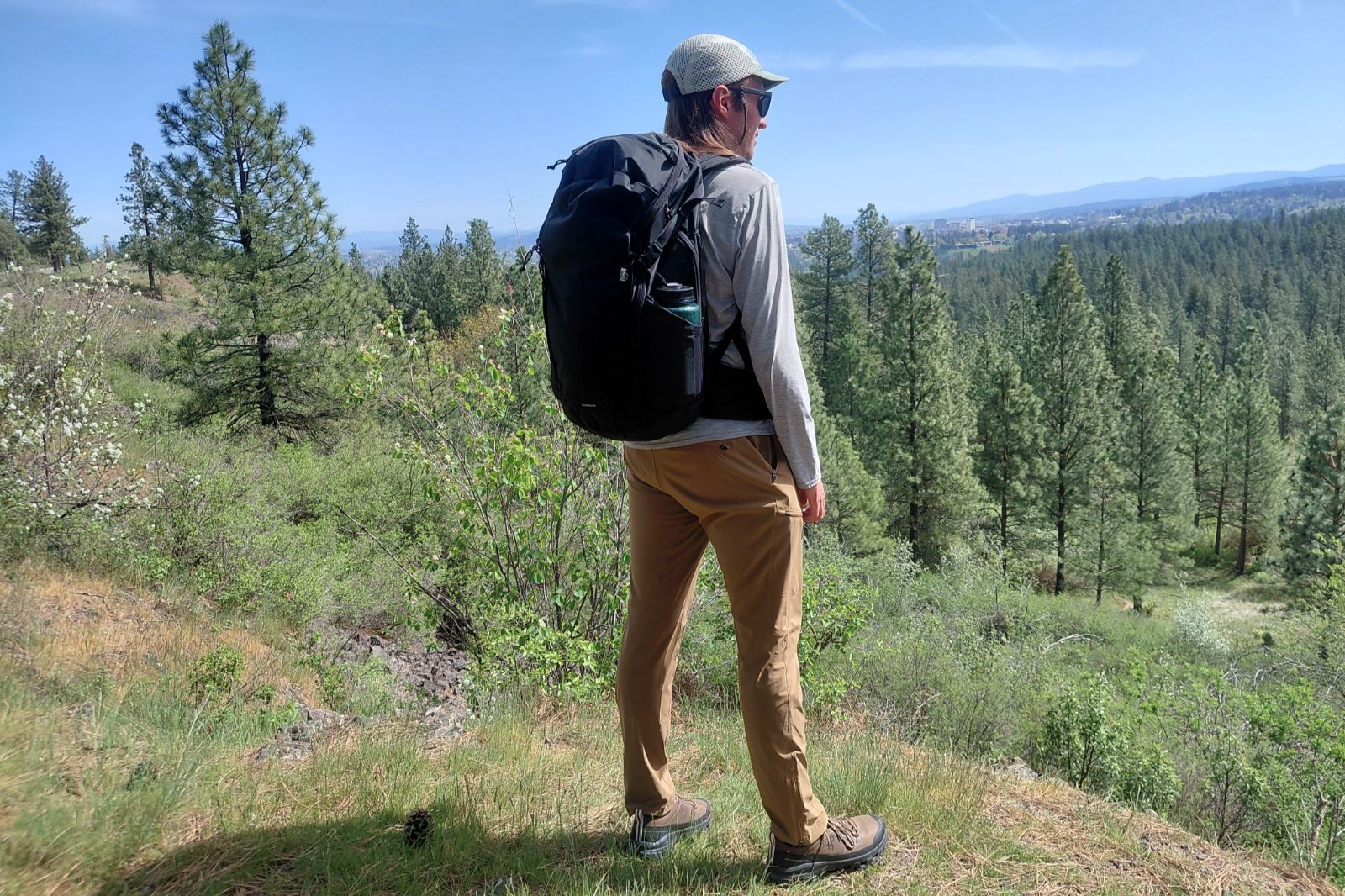 Un hombre con gorra y gafas de sol se encuentra en una colina, contemplando un vasto bosque de pinos bajo un cielo azul claro.