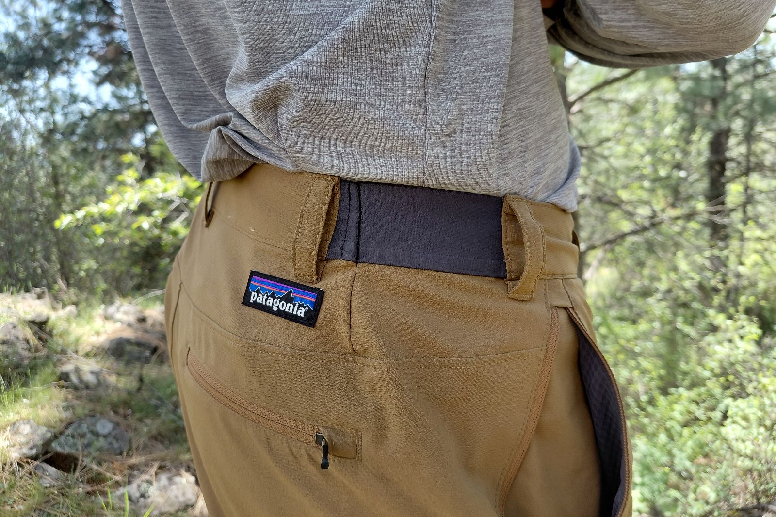 Un primer plano de una persona con pantalones patagónicos marrones y una camisa gris al aire libre en un entorno forestal.