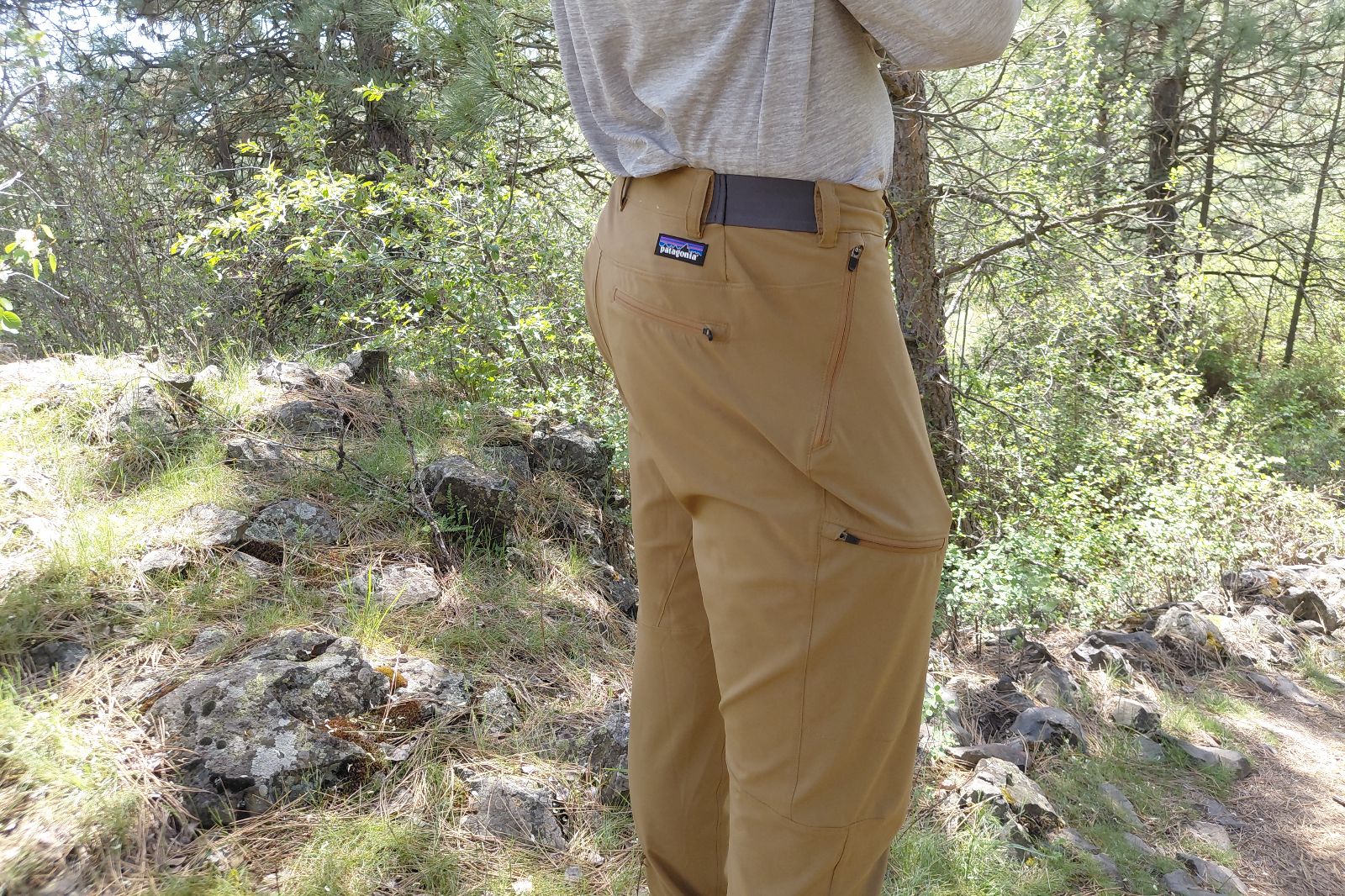 Una persona parada en un sendero forestal con pantalones de senderismo color canela y una camisa gris.