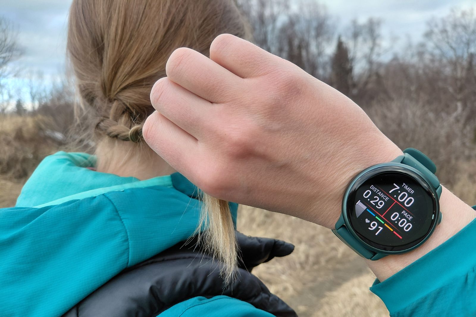 A person wearing a turquoise jacket on a trail shows a smartwatch displaying timer, distance, pace, and heart rate.