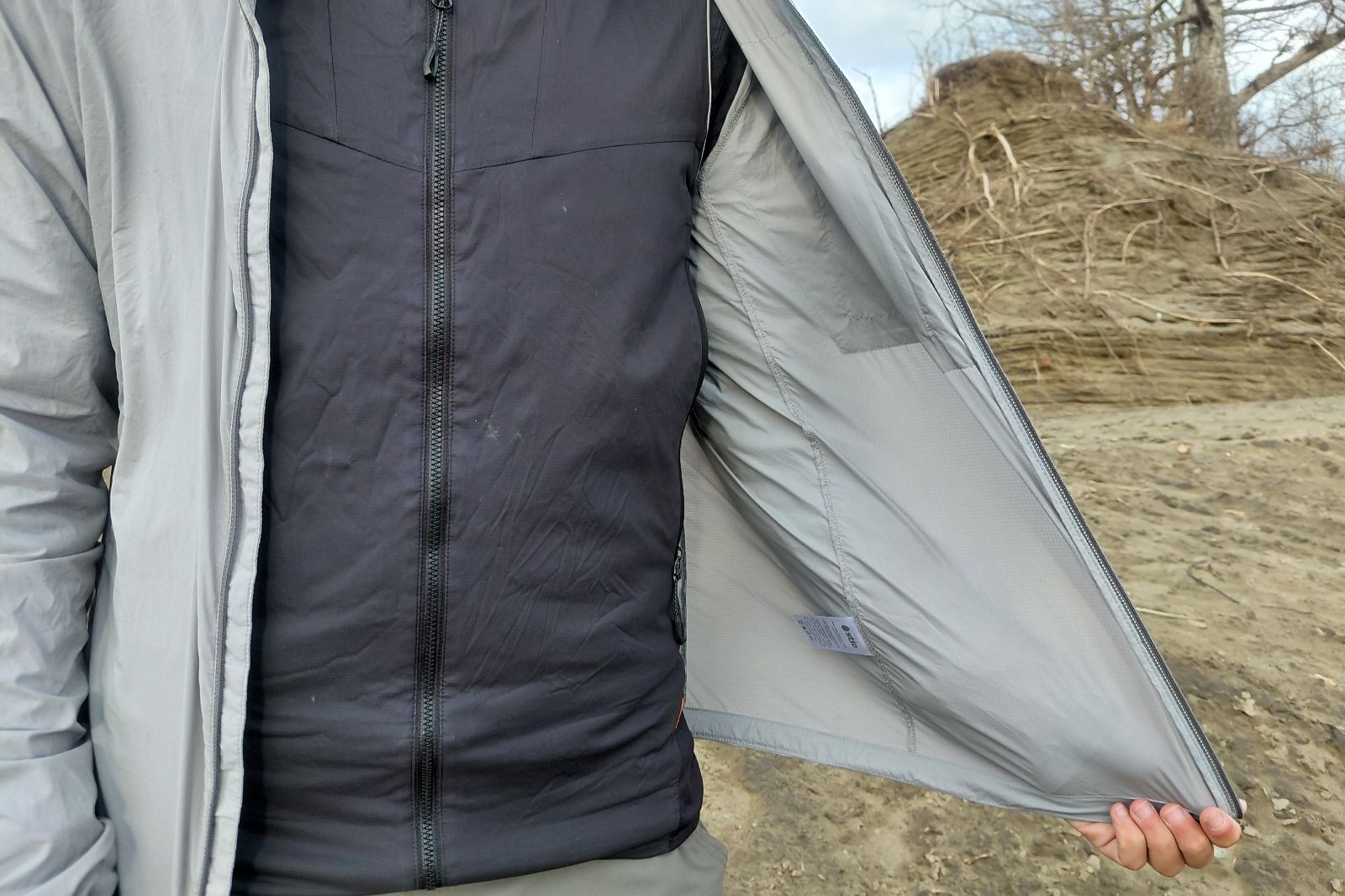 A person in an outdoor setting holds open their light gray jacket, revealing a black zippered inner layer.