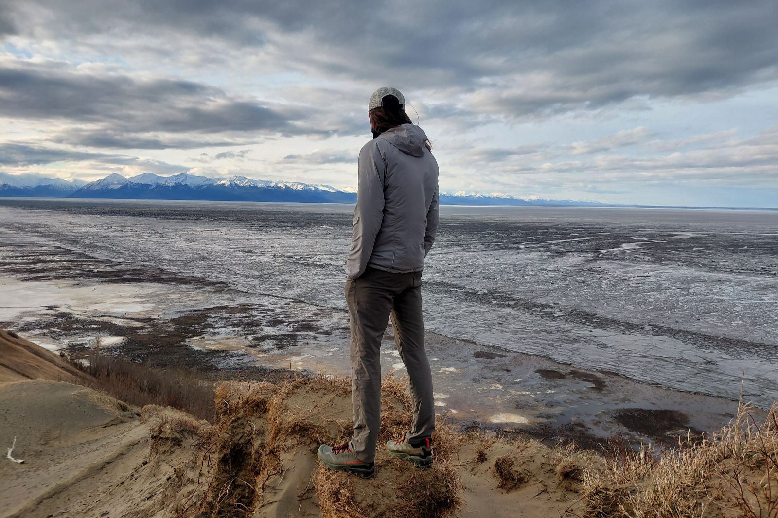 A person stands on a sandy cliff, gazing out over a vast, icy landscape with snow-capped mountains in the distance under a cloudy sky.