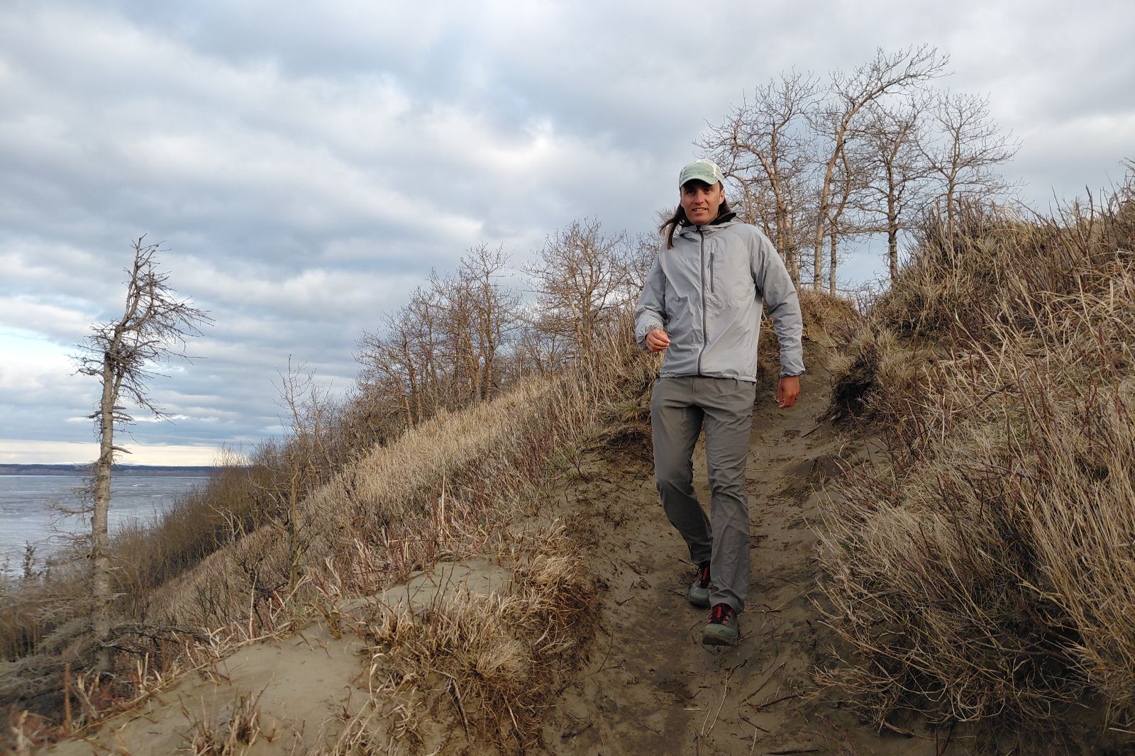 A person in a gray jacket walks down a narrow dirt path in a grassy, barren landscape under a cloudy sky.