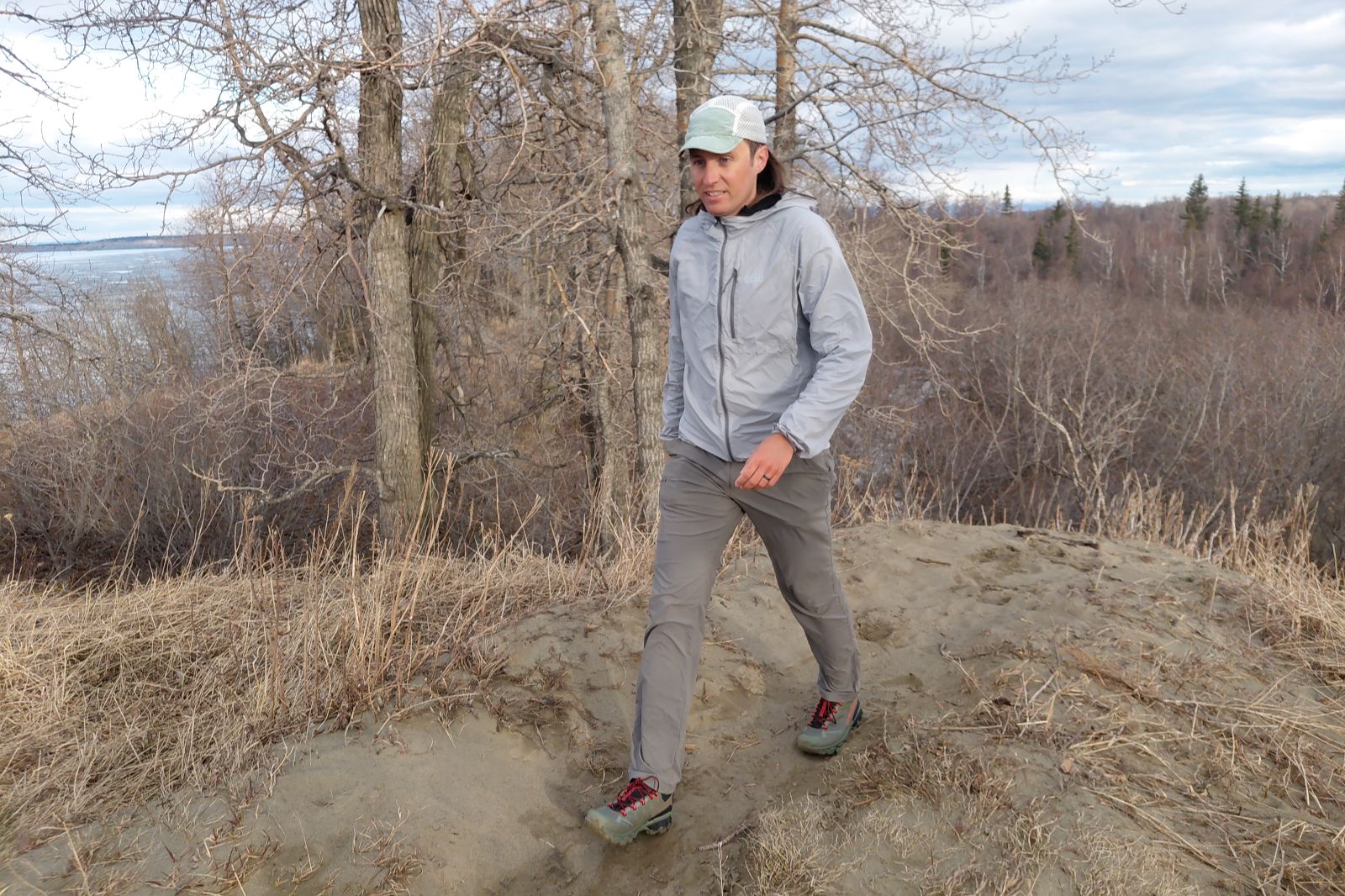 Man in a light jacket and cap walking along a sandy trail, surrounded by leafless trees.