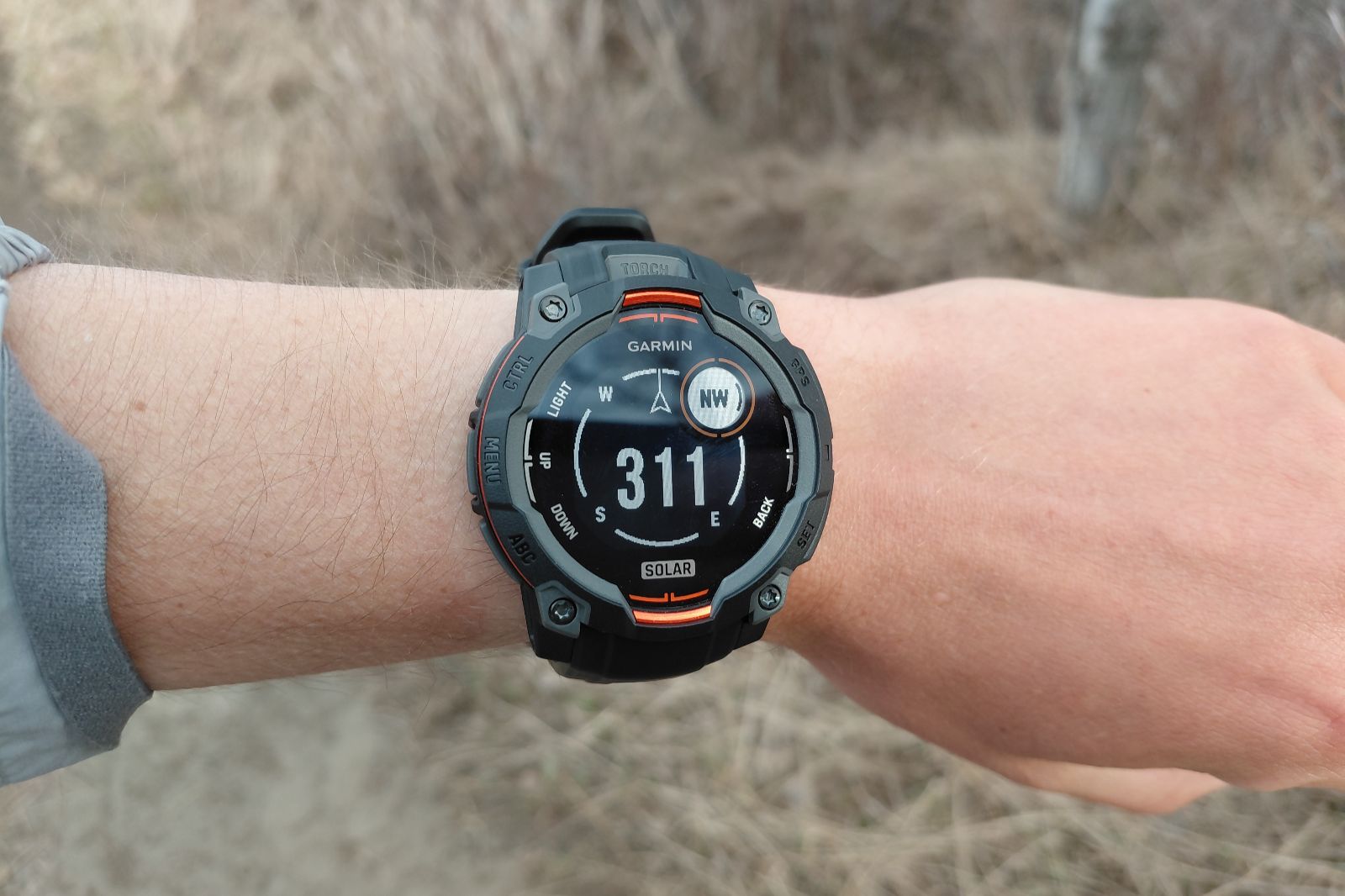 A rugged smartwatch with a compass is worn on the wrist outdoors.