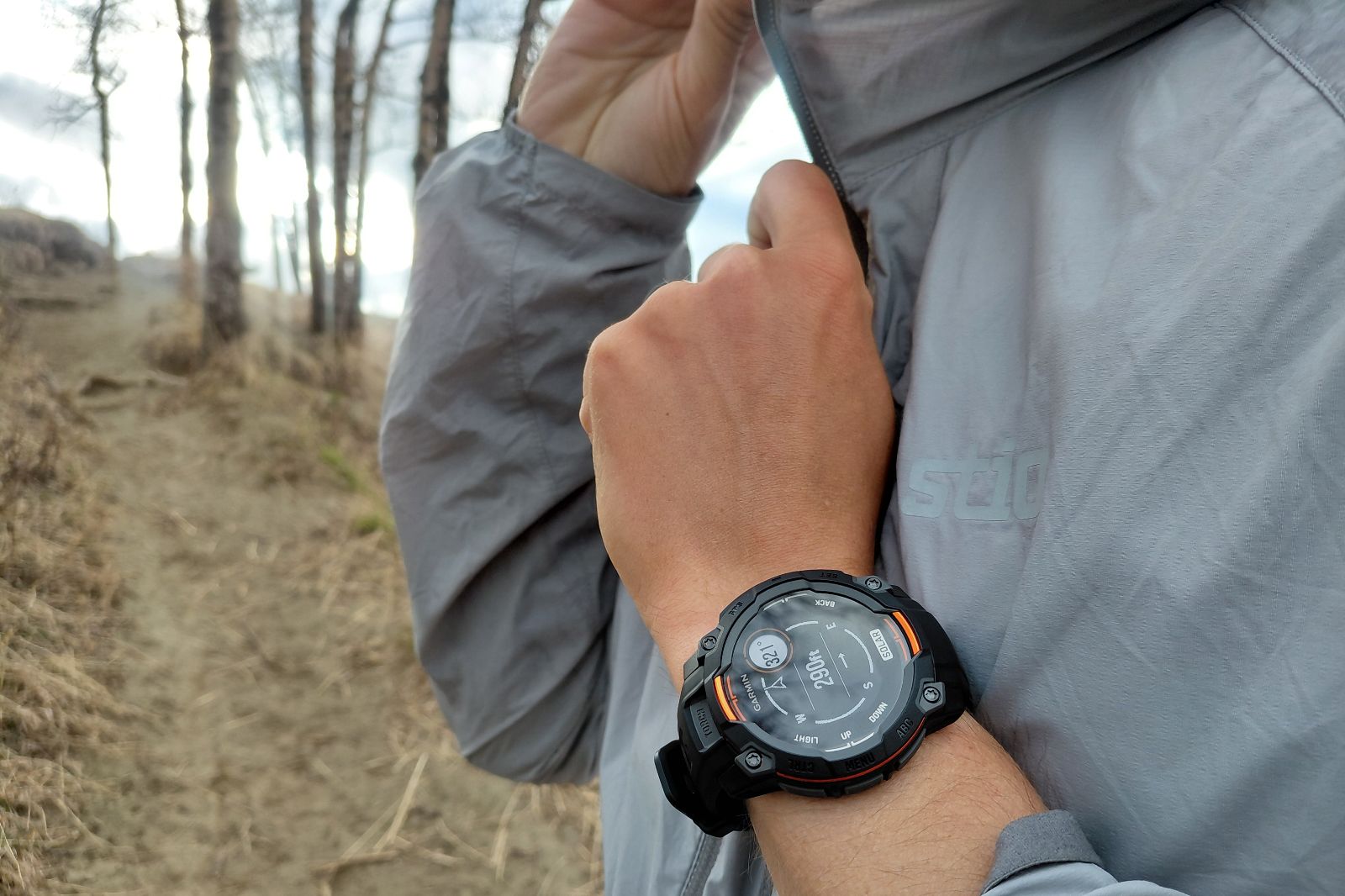 Person wearing a smartwatch and gray jacket zips up while standing on a forest trail.