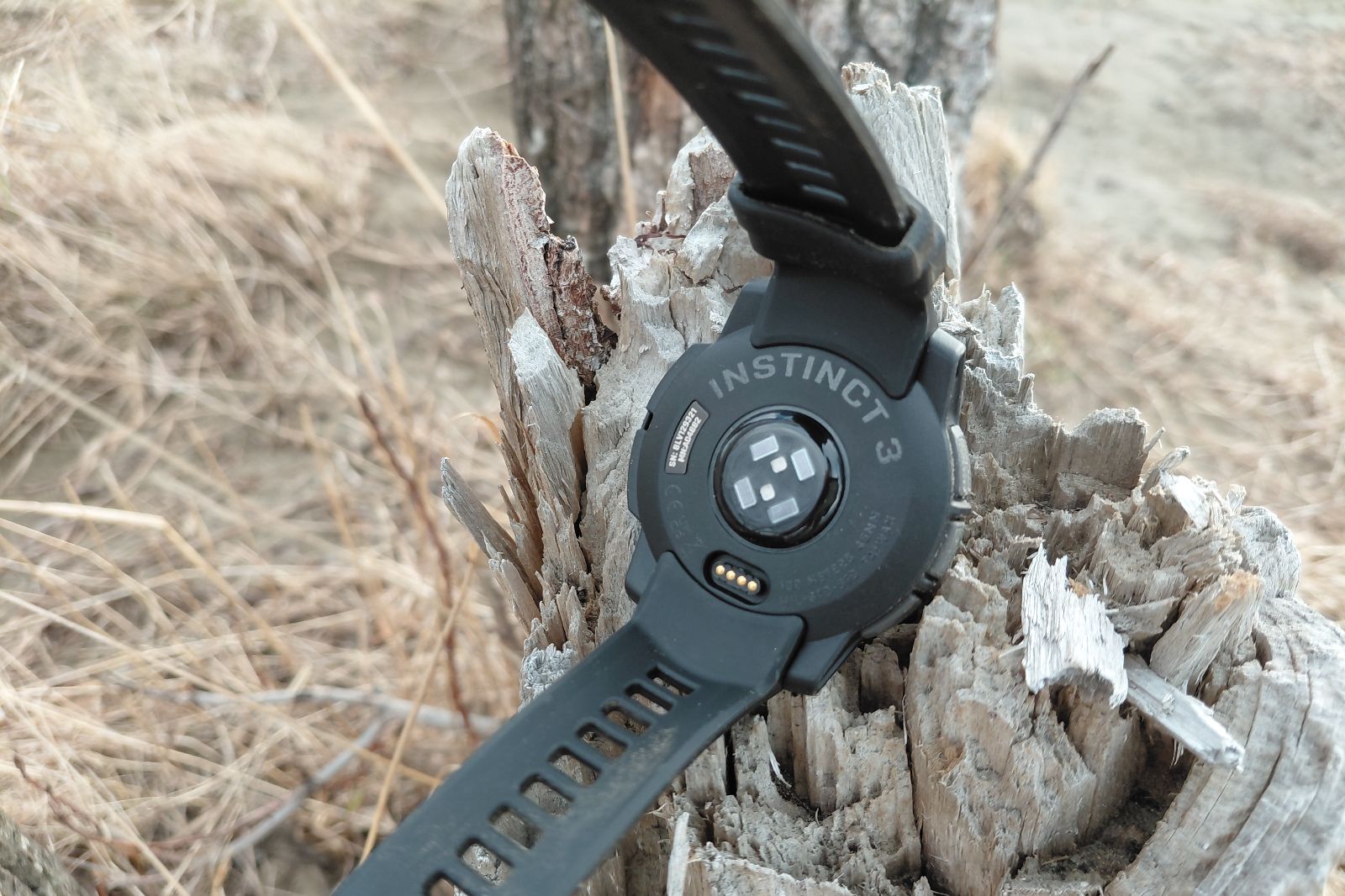 A black wristwatch, labeled "Instinct 3," rests on a weathered tree stump surrounded by dry grass.