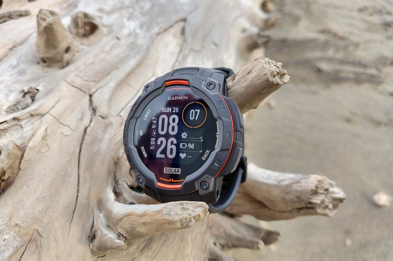Rugged smartwatch with a round face, black and red accents, displaying time and date, mounted on driftwood on a sandy beach.