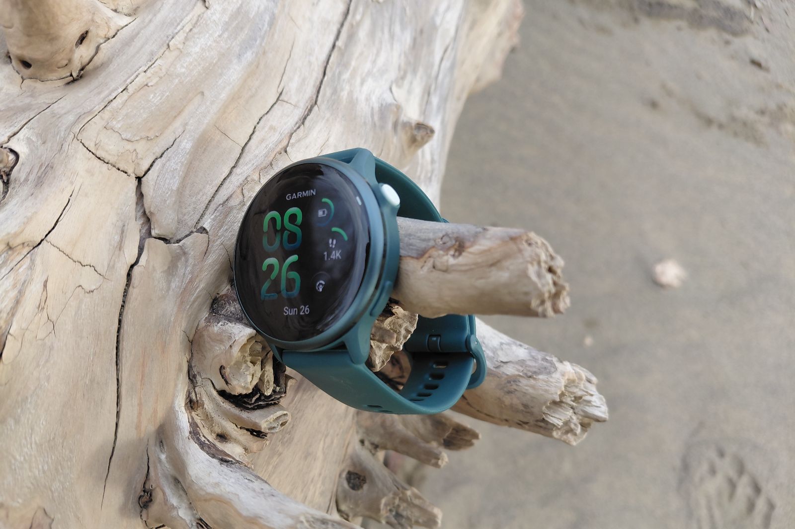 Smartwatch with teal strap displays time against driftwood on a sandy beach.