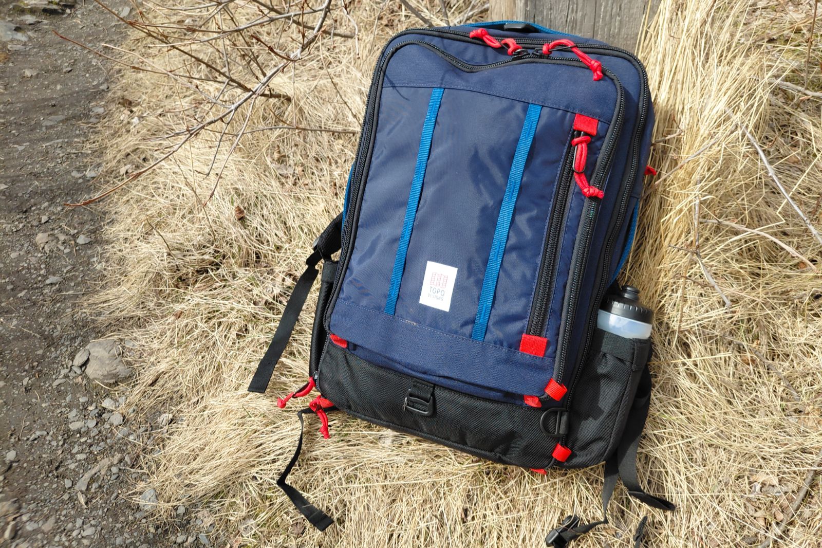 A blue and black backpack with red accents sits on dry grass beside a rocky path.