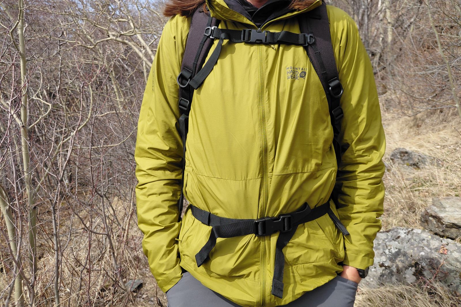 Person wearing a bright green jacket and black backpack, standing on a rocky trail surrounded by leafless trees.