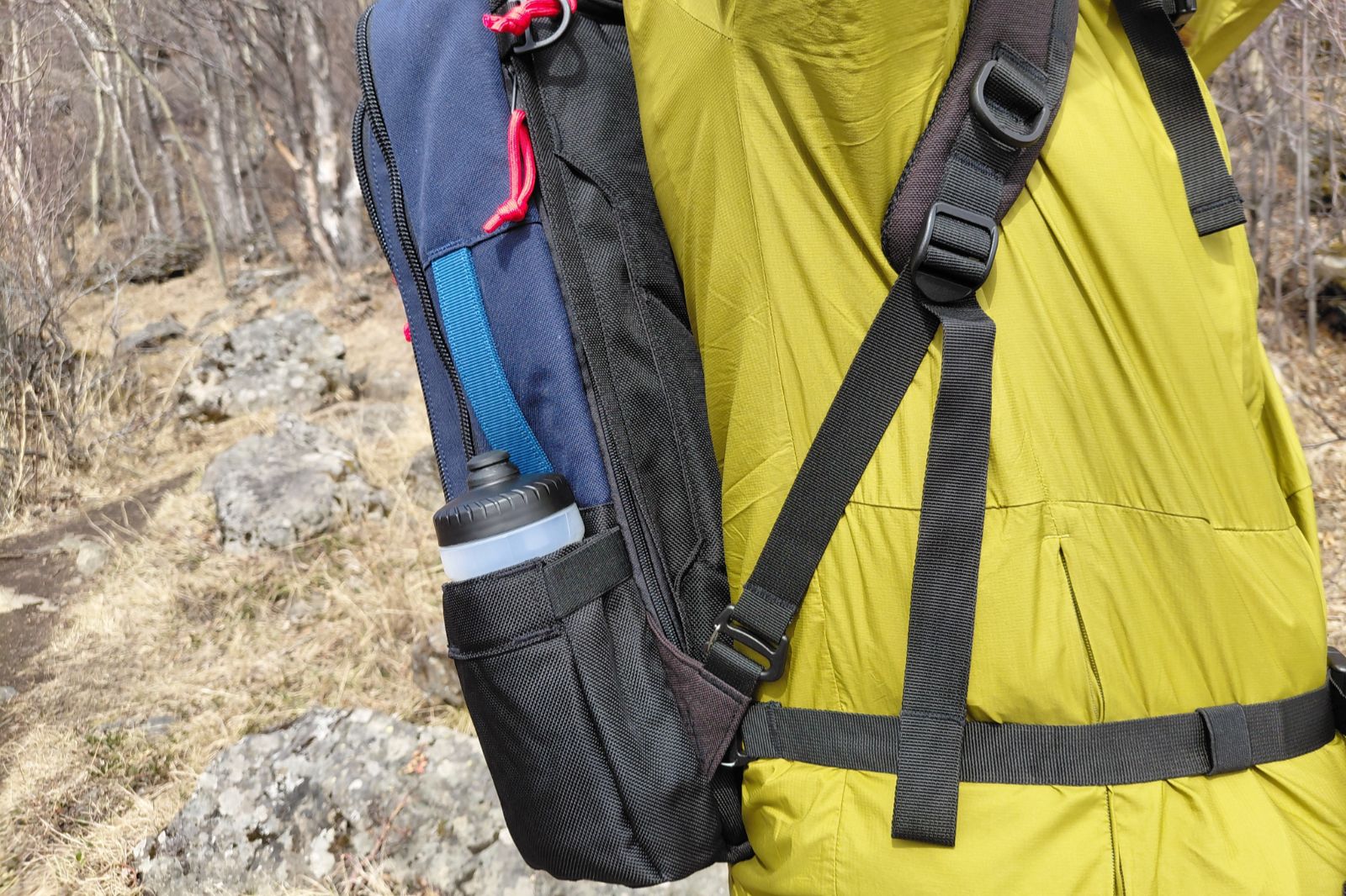 A person wearing a green jacket carries a blue backpack with a water bottle in the side pocket, walking on a rocky forest trail.