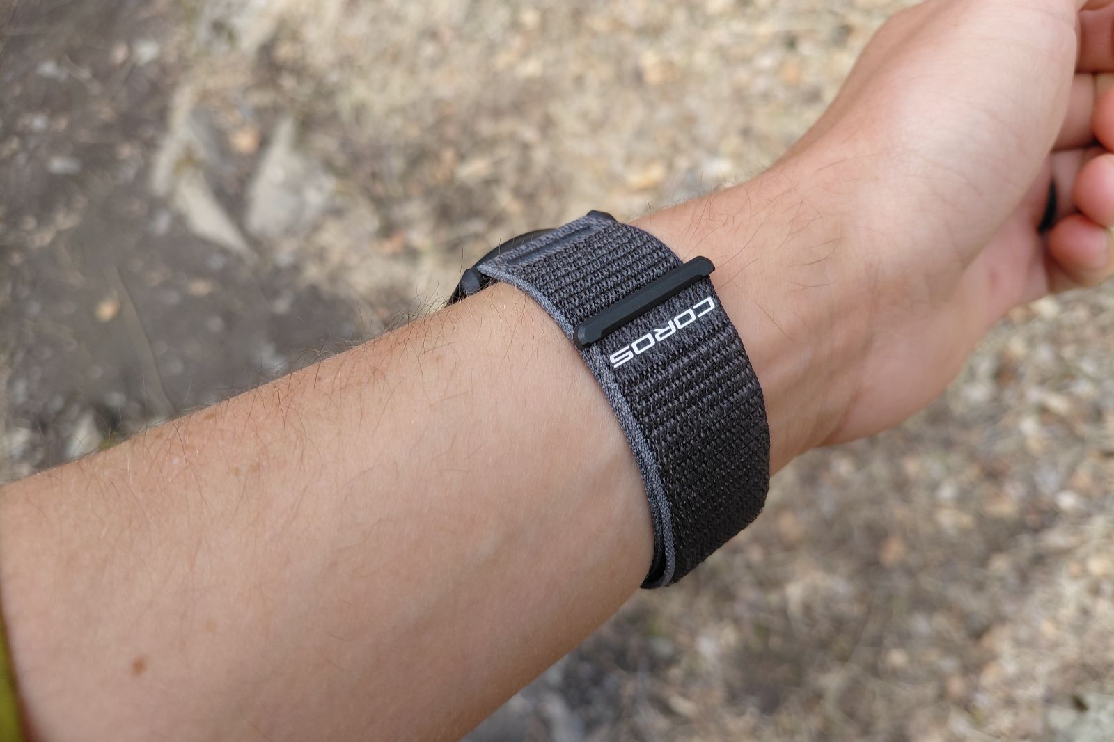 A close-up of a wrist wearing a black Coros nylon strap, set against a blurred outdoor background.