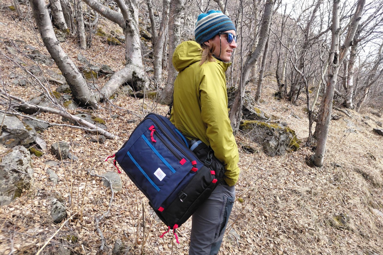 Person in outdoor gear stands in a forested area with bare trees, wearing a green jacket, blue-striped beanie, and sunglasses, carrying a blue travel bag.
