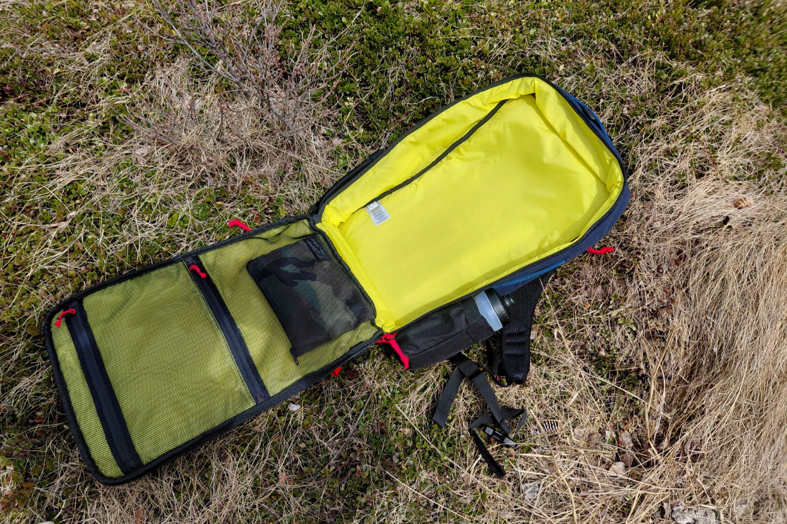 Open outdoor backpack with a bright yellow interior lying on grass. Mesh pockets are visible on one side.