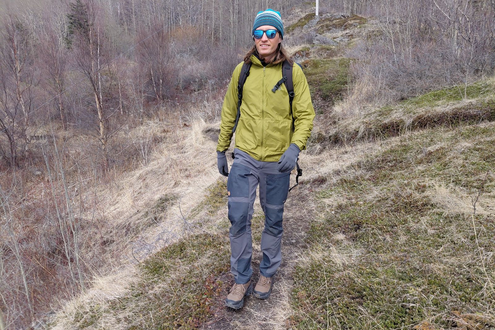 Una persona vestida con equipo para actividades al aire libre, incluida una chaqueta verde y un gorro azul, camina por un sendero cubierto de hierba entre árboles sin hojas.