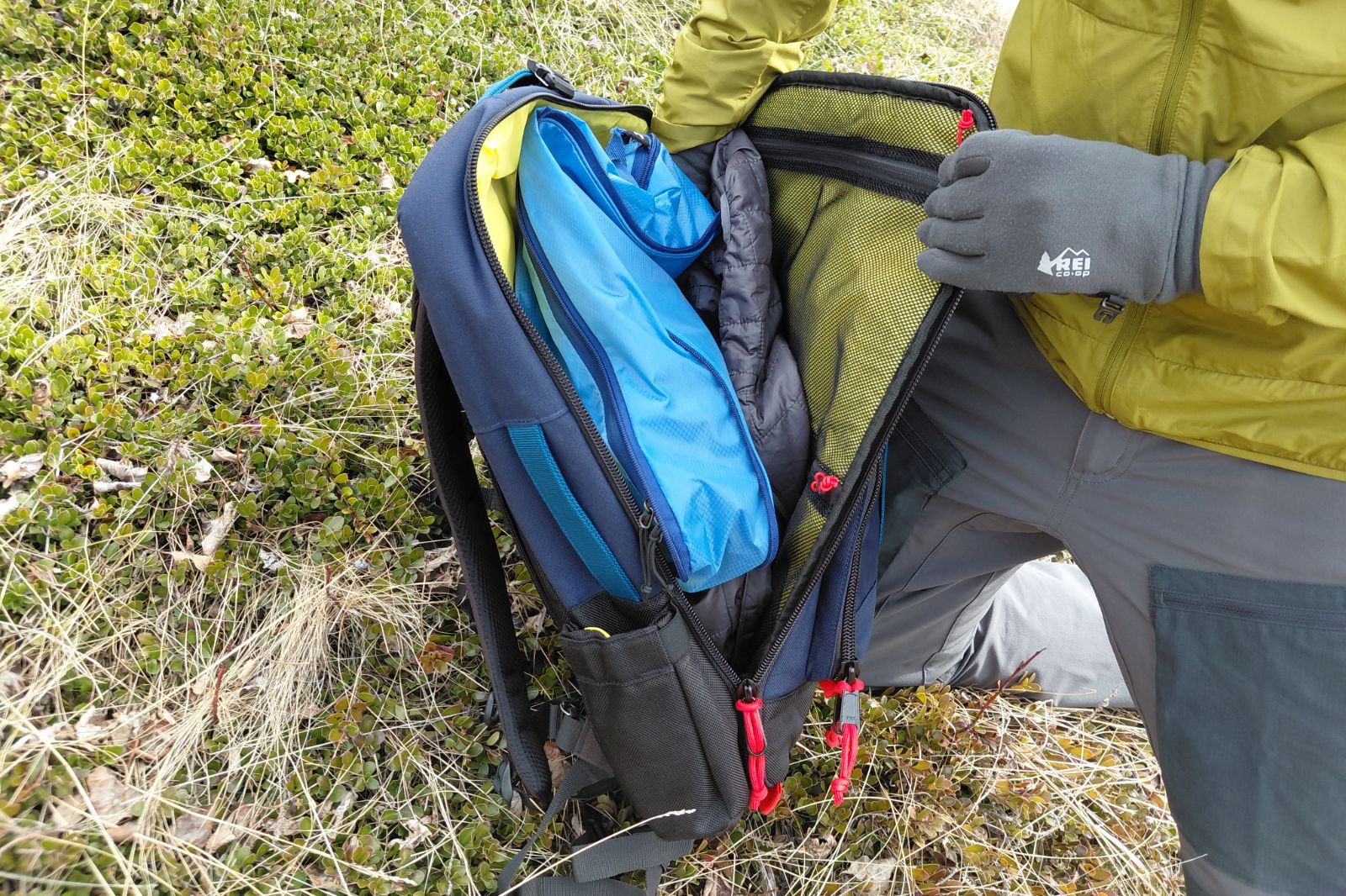 A person in green jacket and gloves kneels on grass, opening a packed travel backpack.