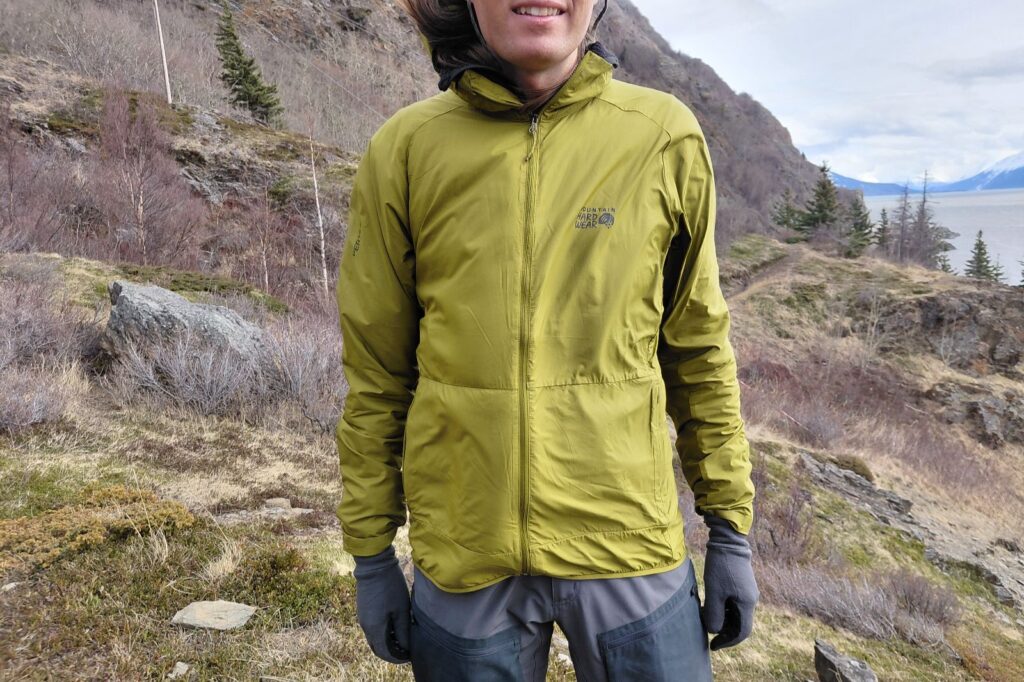 A person stands on an open hillside wearing a green jacket and gray pants.