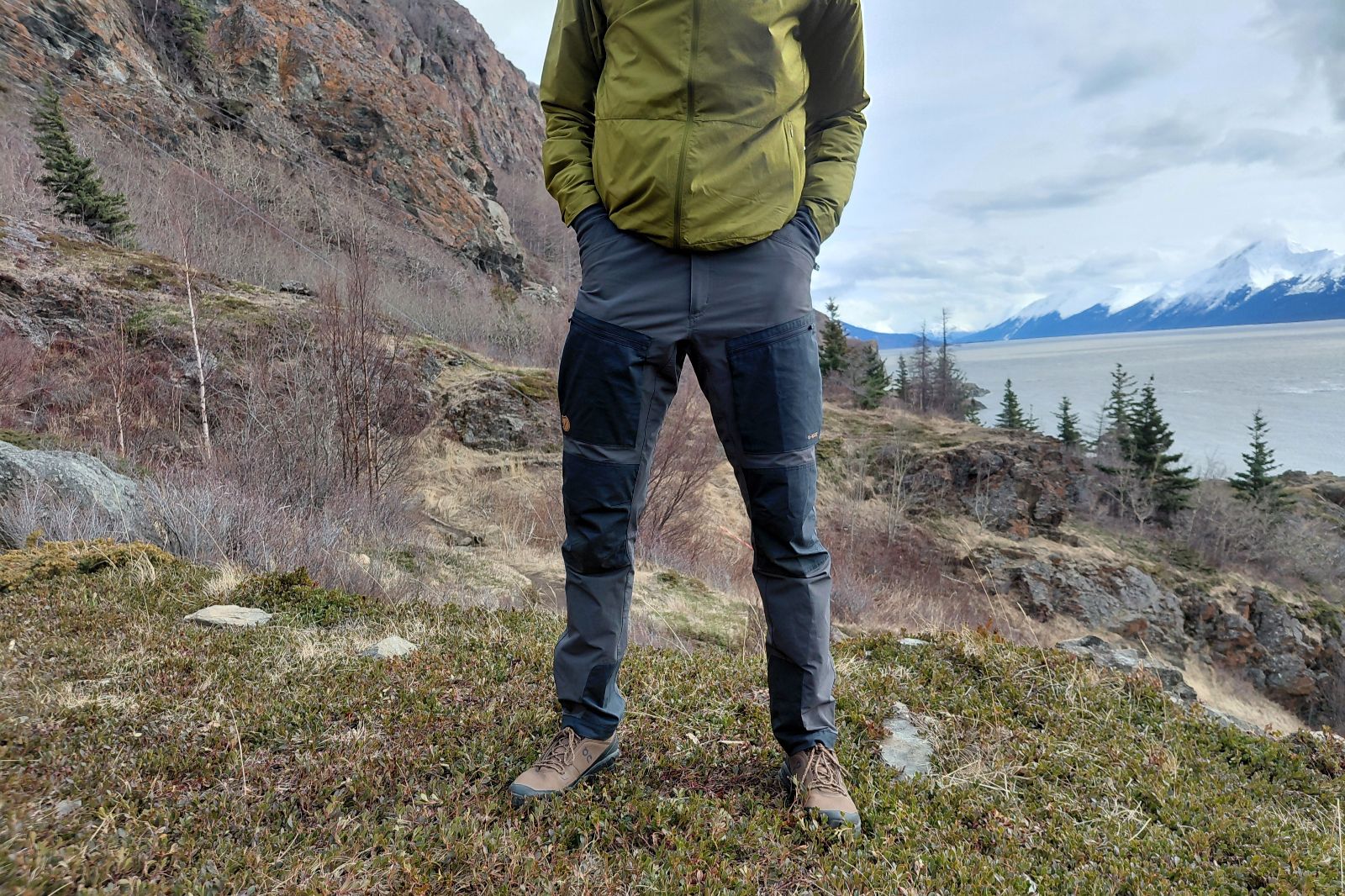 Una persona con una chaqueta verde y pantalones de senderismo se encuentra en un terreno rocoso con las manos en los bolsillos.