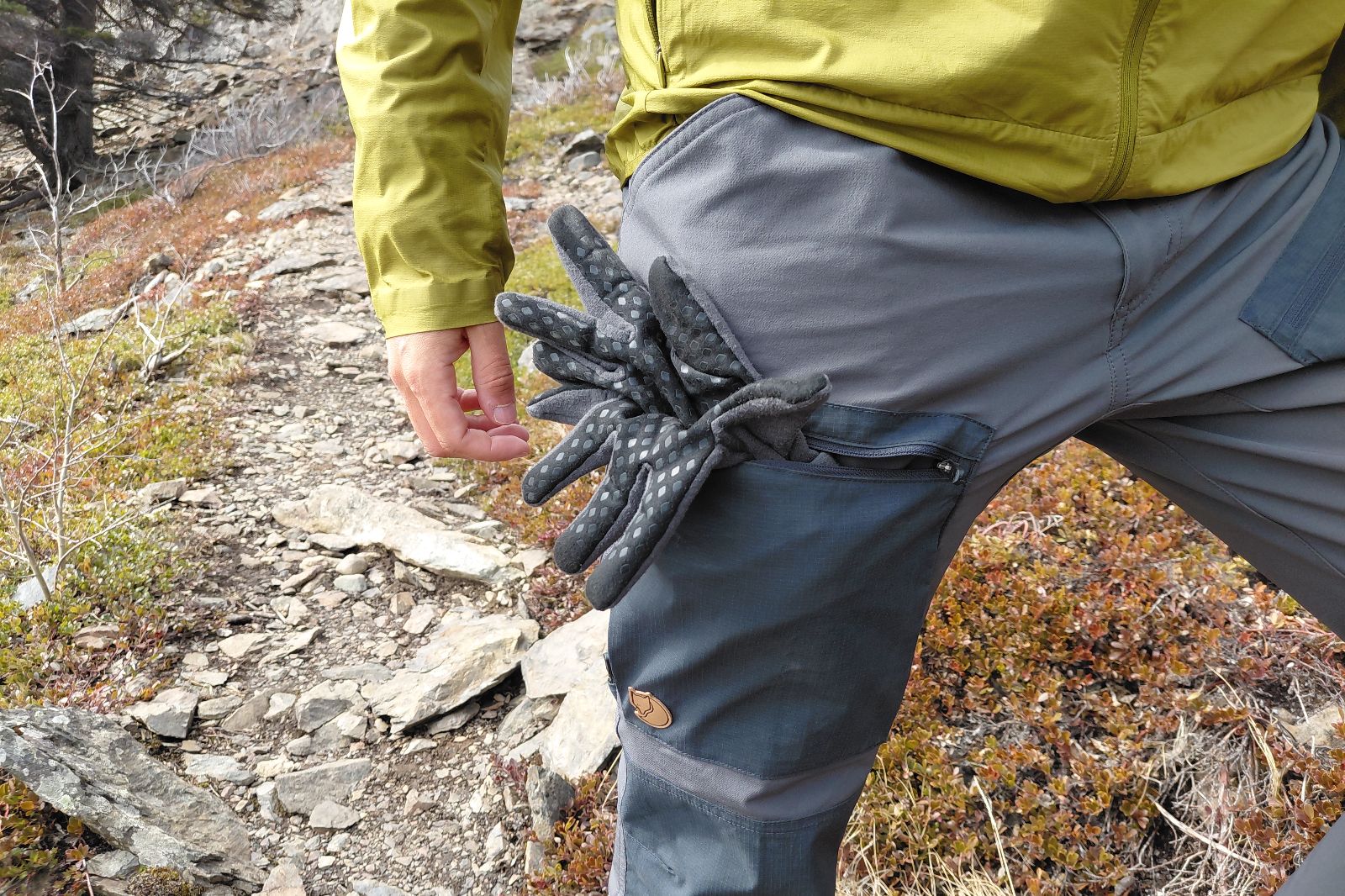 Una persona con equipo de senderismo, una chaqueta verde y pantalones grises con guantes metidos en un bolsillo, camina por un sendero de montaña rocosa.