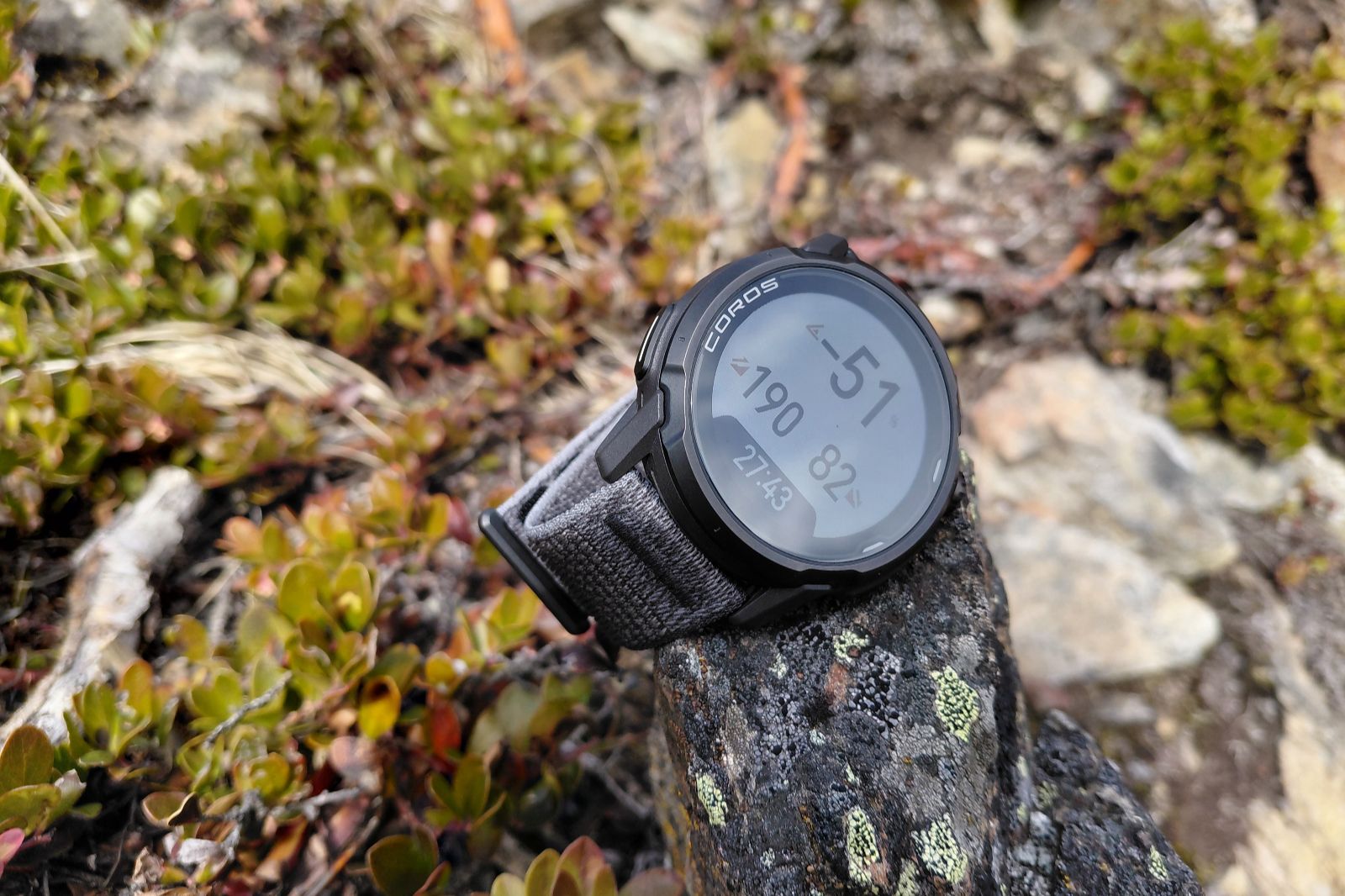 A black GPS watch with a fabric band rests on a moss-covered rock.