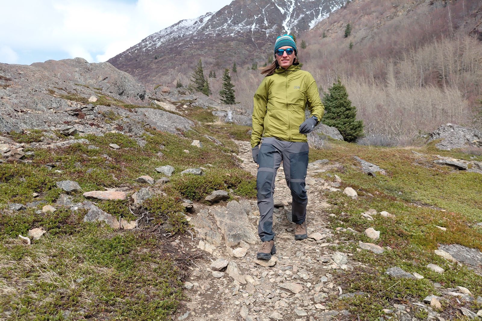 A person hiking on a rocky trail in a mountainous area, wearing a green jacket and blue hat.