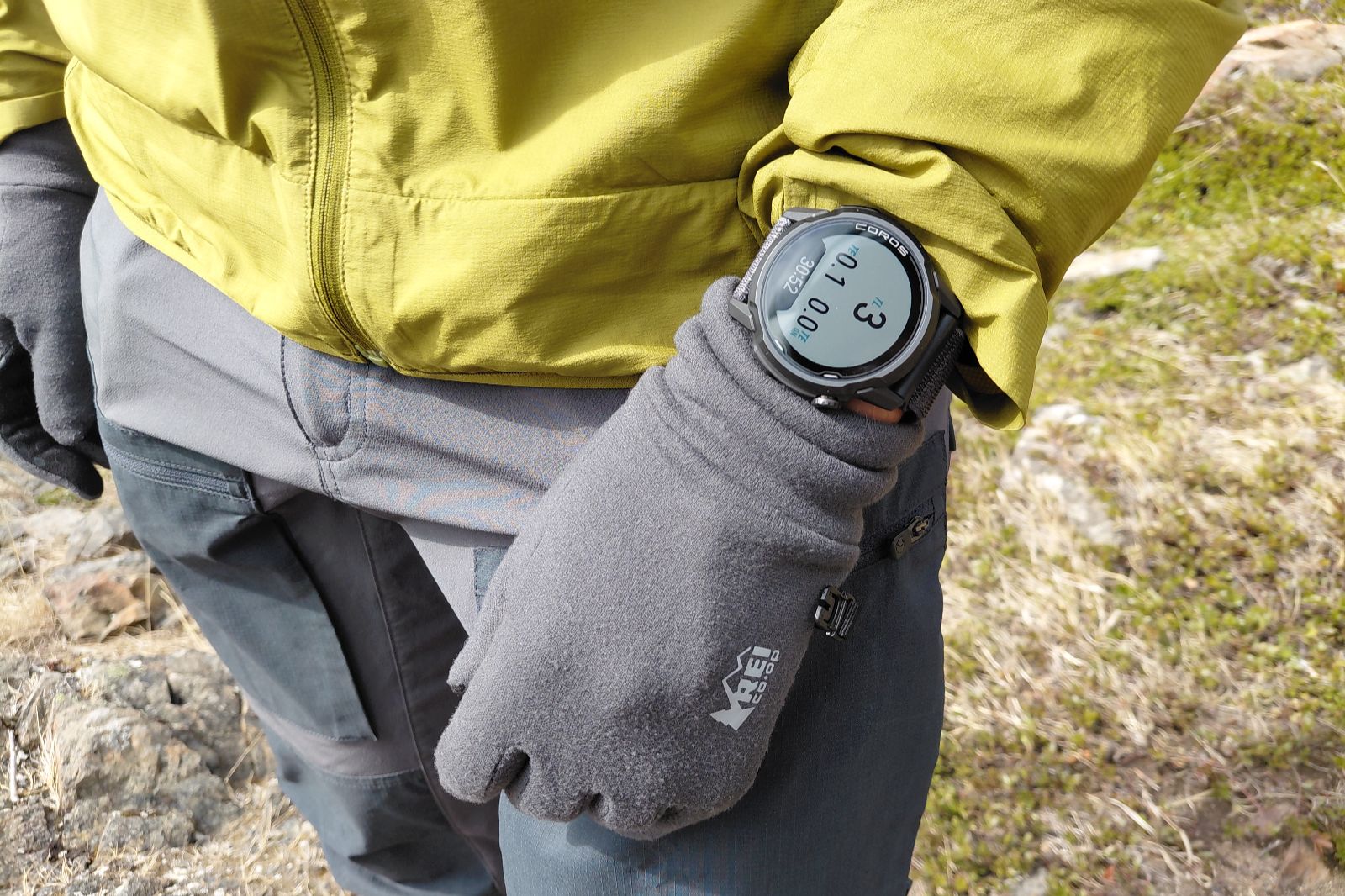 A person in a yellow jacket and gray gloves checks an outdoor watch with a digital display.