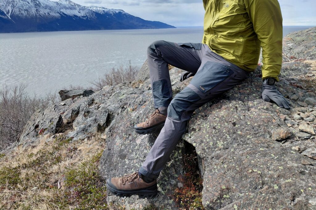 A person in an olive jacket and gray pants sits on a mossy rock, overlooking the ocean with snow-capped mountains in the background.