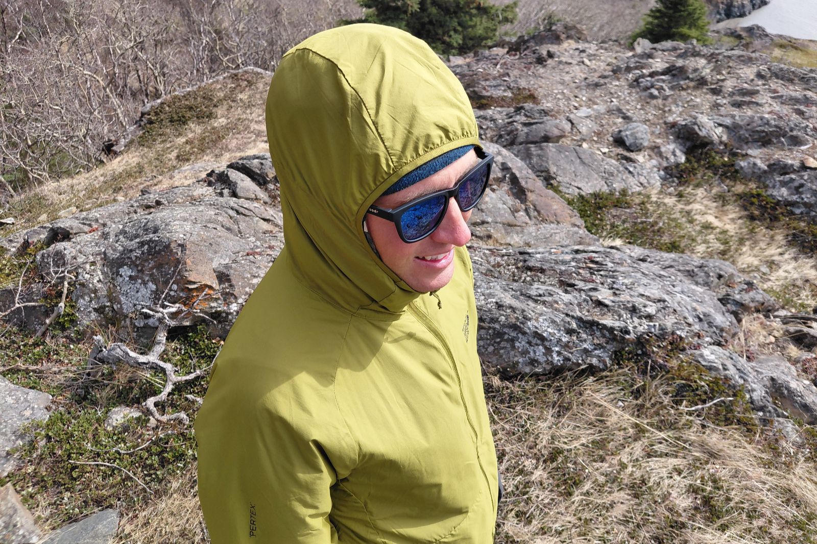 A person in a green hooded jacket and sunglasses stands on rocky terrain, smiling.