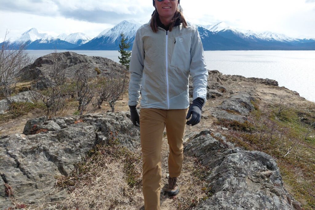 A person wearing a light jacket and brown pants hikes on a rocky trail above the ocean.