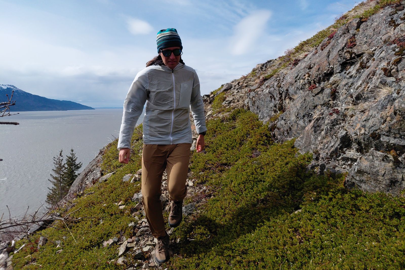 Person wearing a jacket and beanie walks along a rocky, grassy hillside near a vast, cloudy seascape.