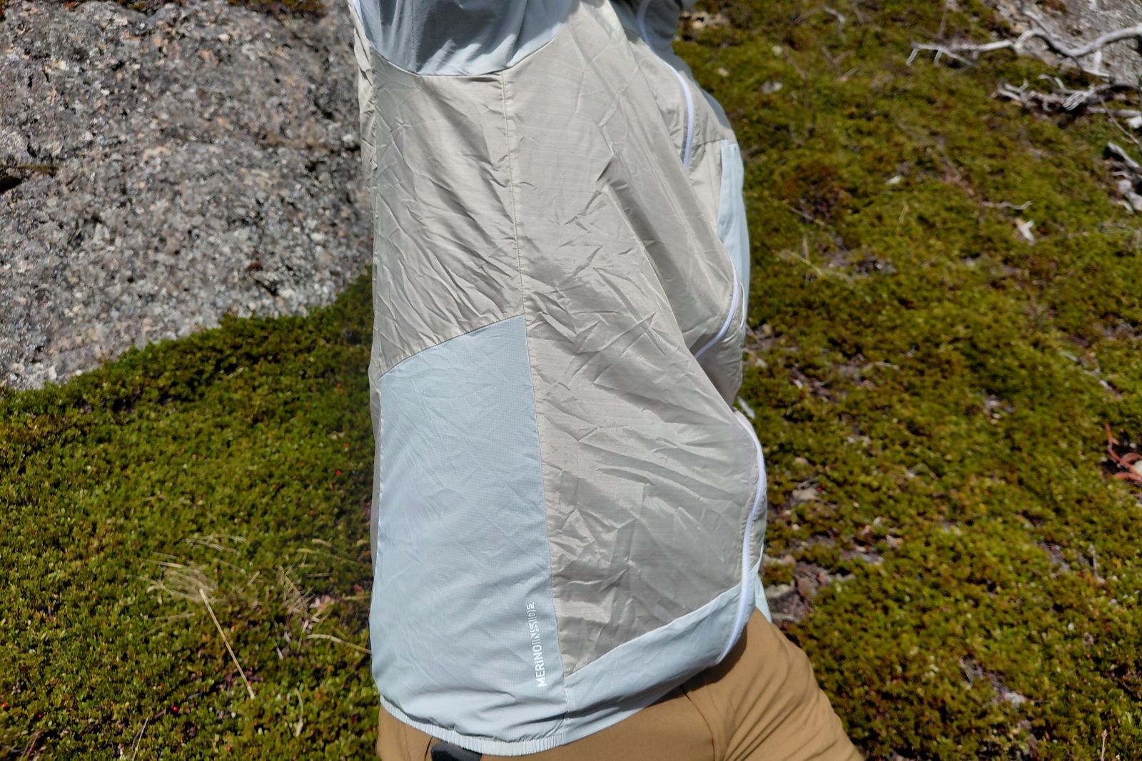 A person in a light hiking jacket stands with one arm up, showing the side panels.