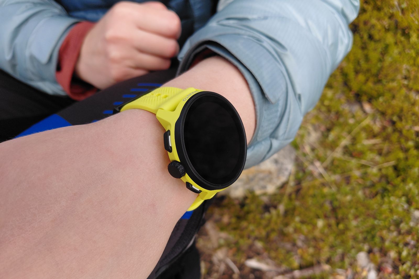 Close-up of a person wearing a smartwatch with a bright yellow wristband, set against an outdoor mossy ground