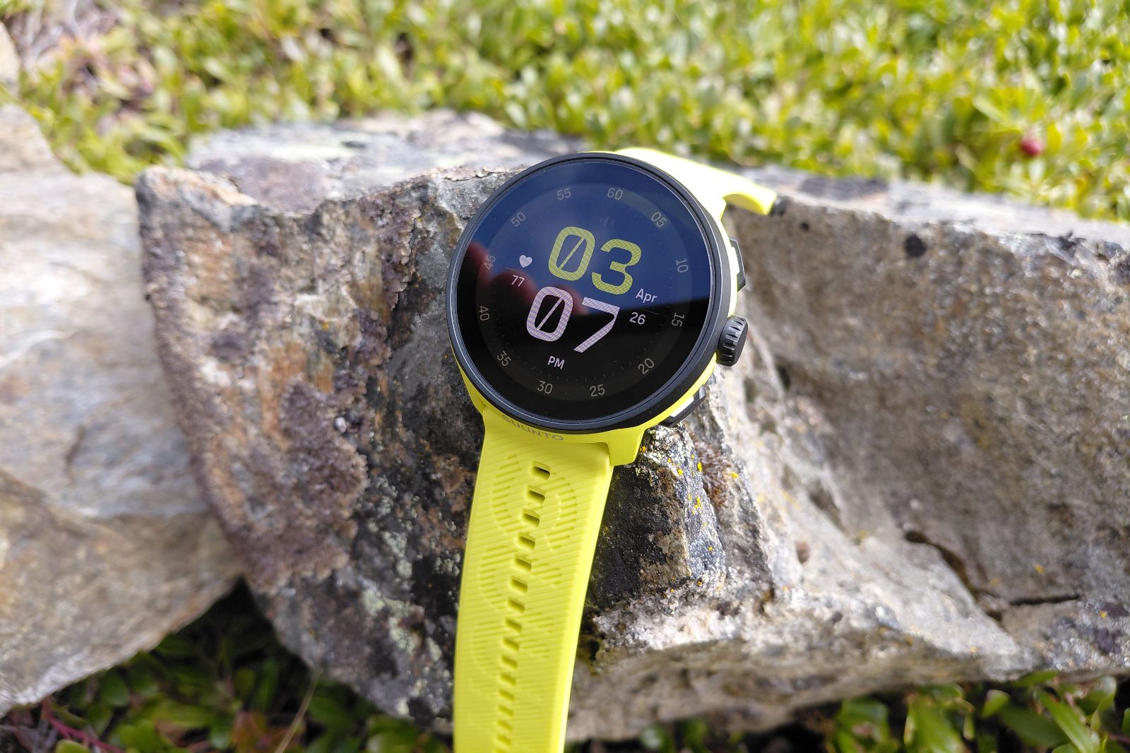 Yellow smartwatch with a round face displaying time, resting on a rock.