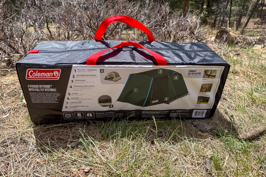 The Coleman Skydome 4 tent packed in its rectangular carry bag with red handles, resting on dry grass.